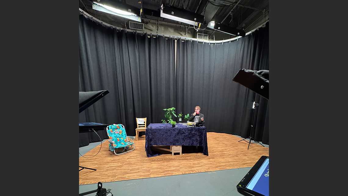 Actor on stage, sitting behind a table, with a view of technical equipment on the sides