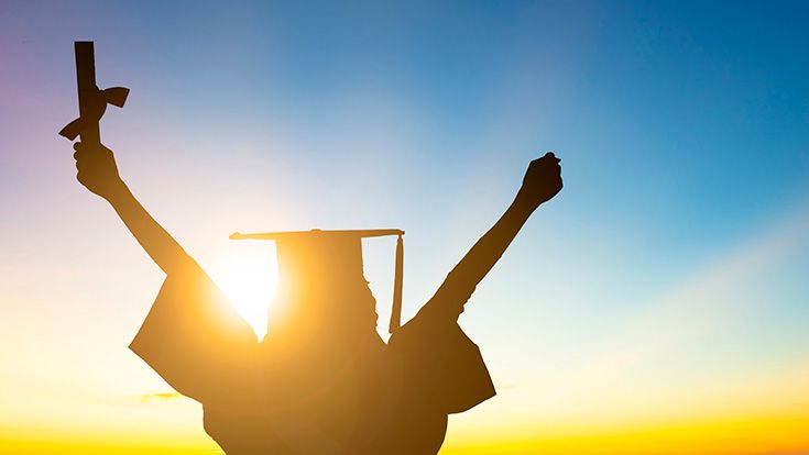 A graduate looking into a sunset holding arms in the air with a diploma in hand